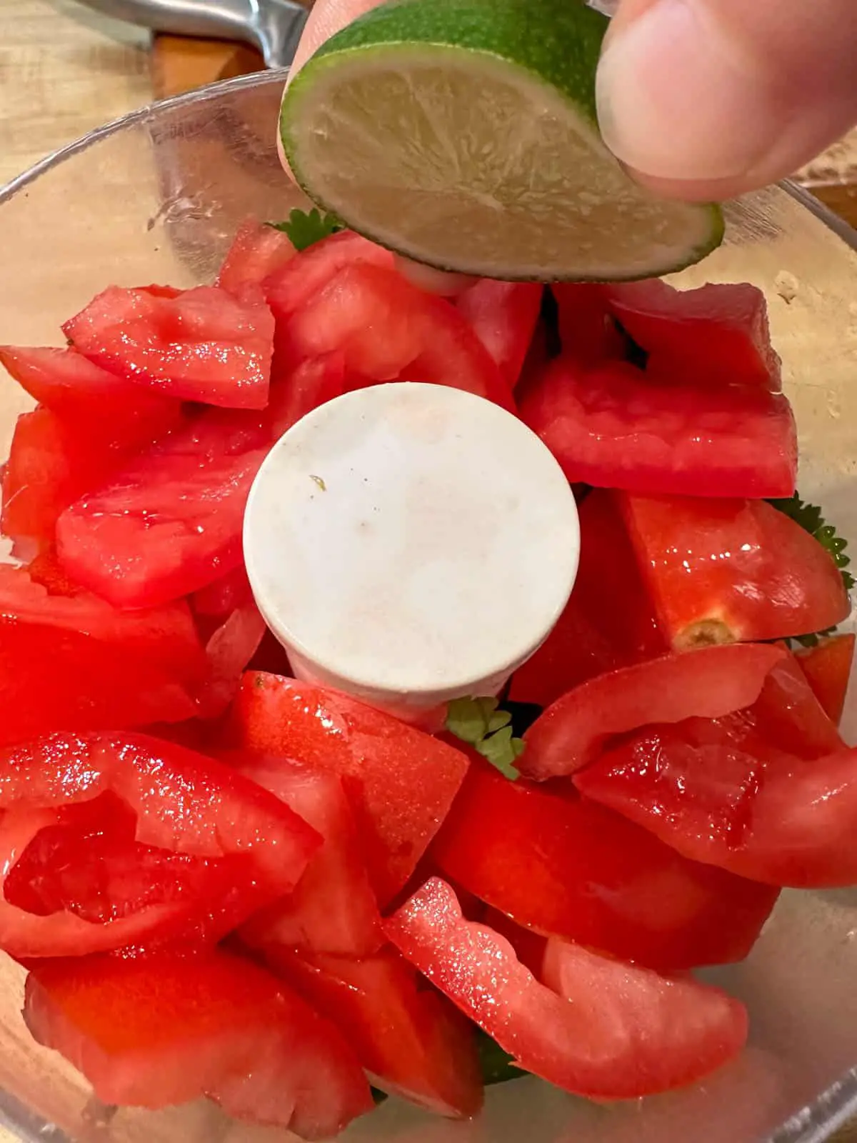 A food processor containing ingredients for the salsa with chopped tomatoes on the top. There is someone holding a lime slice over the food processor.