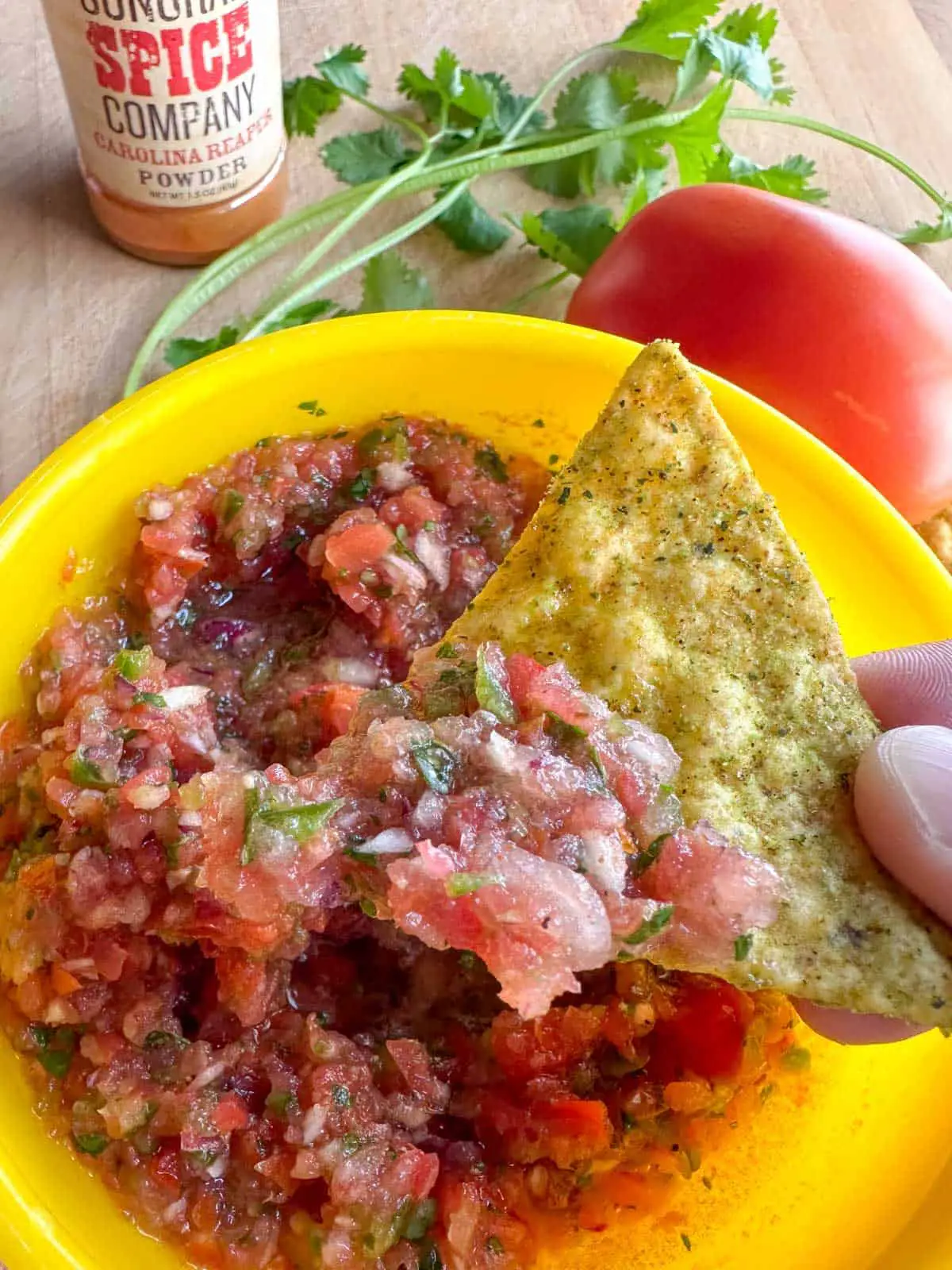 Carolina Reaper salsa in a yellow bowl with cilantro, and a tomato surrounding the bowl. There is a bottle of Carolina Reaper powder in the background and a person holding a tortilla chip which has been dipped into the salsa.