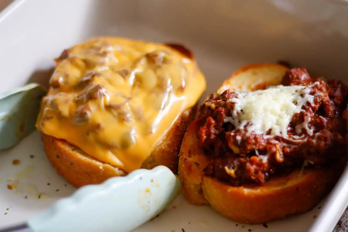 A white casserole dish with 2 pieces of Texas Toast sloppy joes one topped with melted cheddar and the other with melted American cheese. There is a pair of blue tongs in the dish.