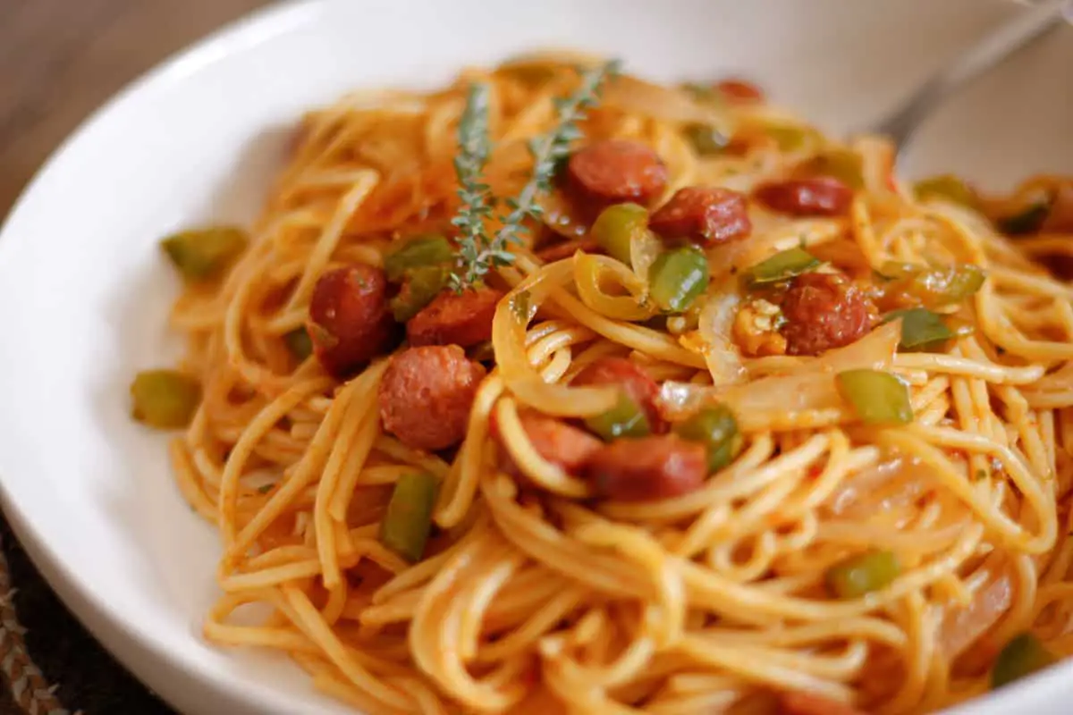 A white plate containing Haitian spaghetti which is spaghetti with hot dogs, sliced onions, and diced green bell peppers. There is a spoon resting in the bowl and thyme sprigs garnishing the dish.