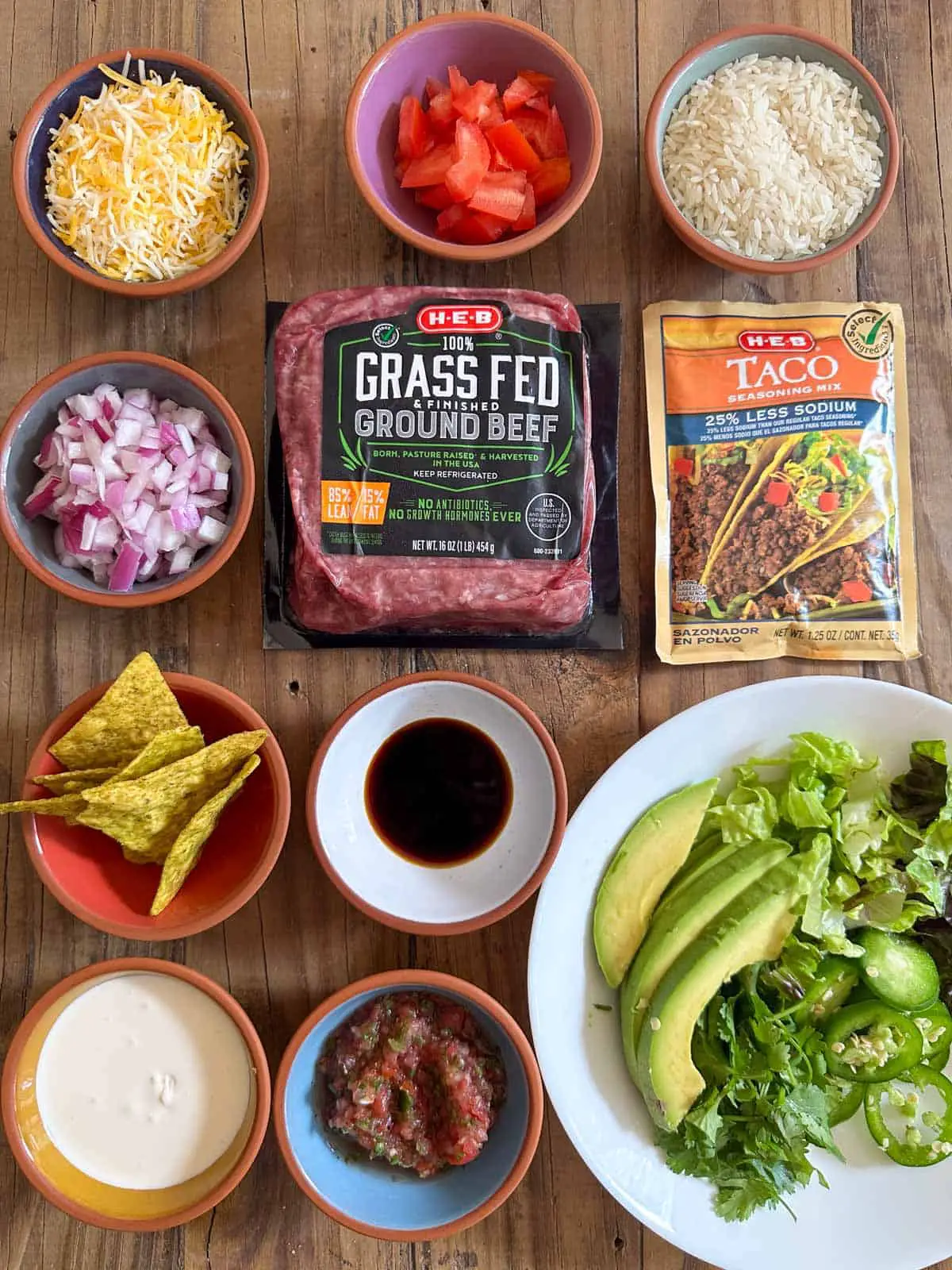 Bowls containing shredded cheese, diced tomato, rice, diced onion, tortilla chips, soy sauce, sour cream, salsa, and lettuce, avocado, cilantro and sliced jalapenos. There is also a pack of Grass Fed Ground beef and Taco seasoning mix.