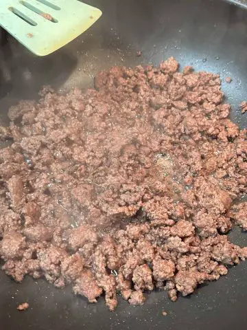 Browned ground beef and its fat in a wok with a blue slotted turner resting on the rim of the wok.