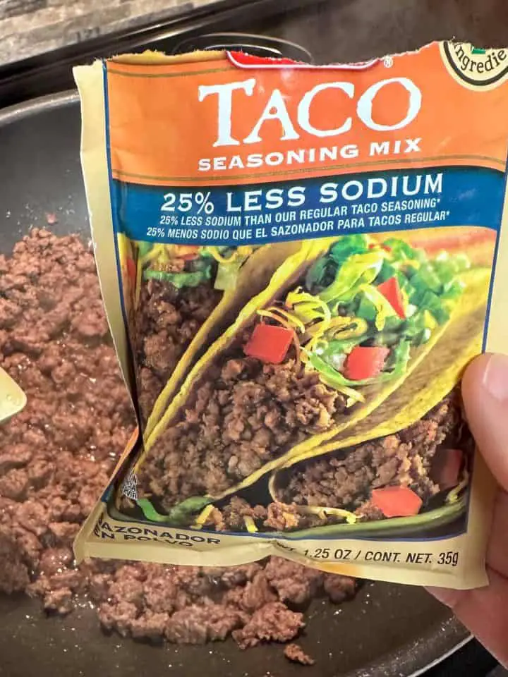 A wok with browned ground beef in the background and a packet of Taco Seasoning Mix being held by someone's hand in the foreground.