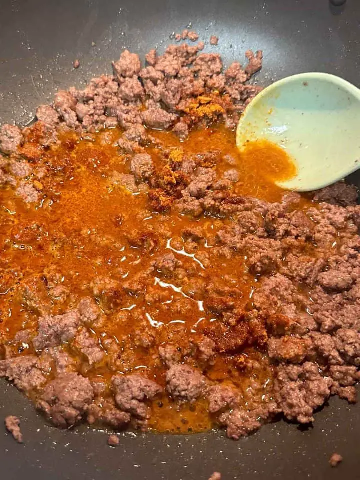 Browned ground beef with taco seasoning and water added to it in a wok with a blue silicone spoon resting in the wok.