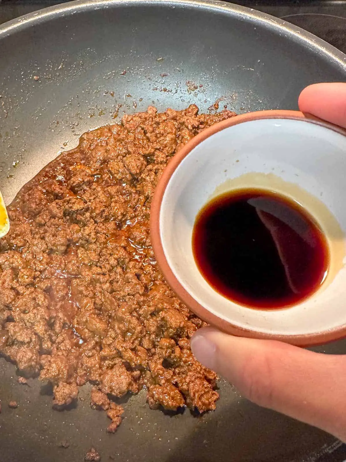 Browned ground beef cooked with taco seasoning in a wok in the background and a small bowl with soy sauce being held by someone in the foreground.