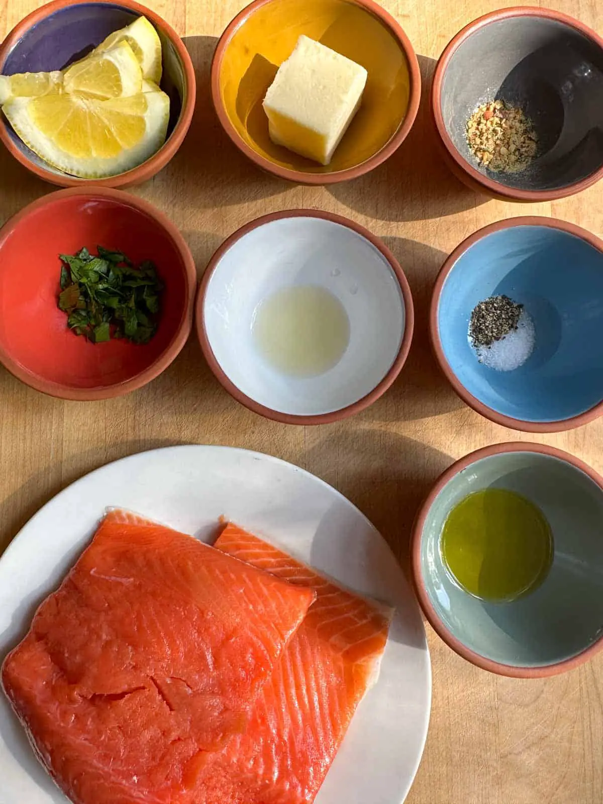 A white plate with raw salmon fillets and small bowls containing slices of lemon, butter, garlic blend seasoning, minced parsley, lemon juice, salt and pepper, and olive oil.