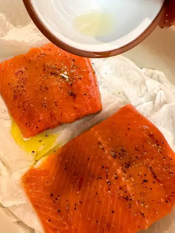 Raw salmon fillets seasoned with salt and pepper atop damp paper towels. A person is holding a small bowl with lemon juice above the fillets.