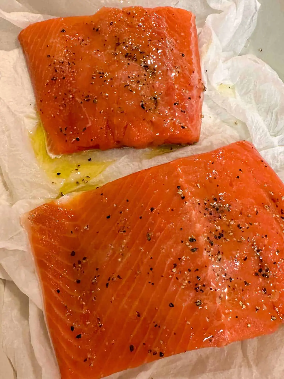 Raw salmon fillets seasoned with salt and pepper and drizzled with olive oil atop paper towels.