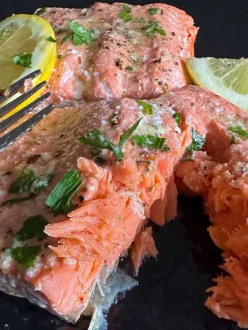 Cooked salmon fillets seasoned with salt and pepper and garnished with Italian parsley and lemon slices. There is a fork on the side, and one of the fillets has been cut into.