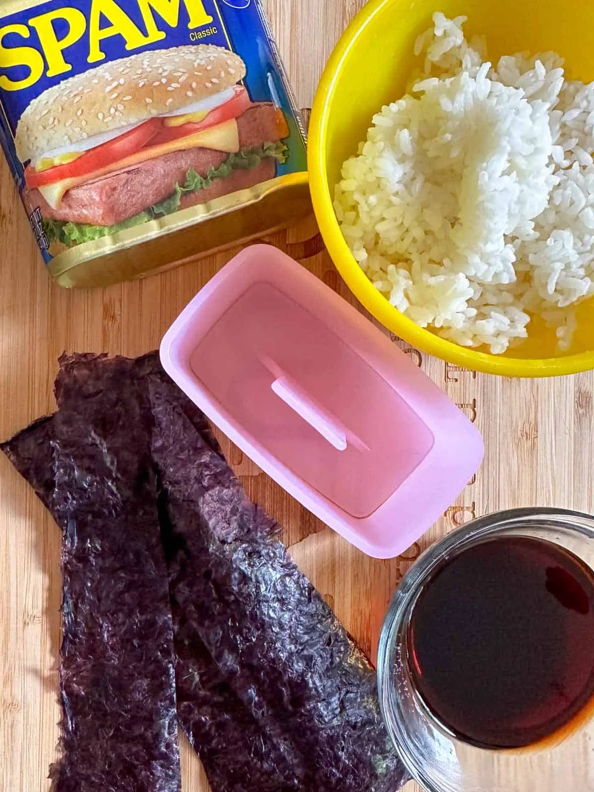 A can of Spam, a yellow bowl with cooked rice, a pink musubi maker, strips of nori, and a glass bowl with soy sauce.