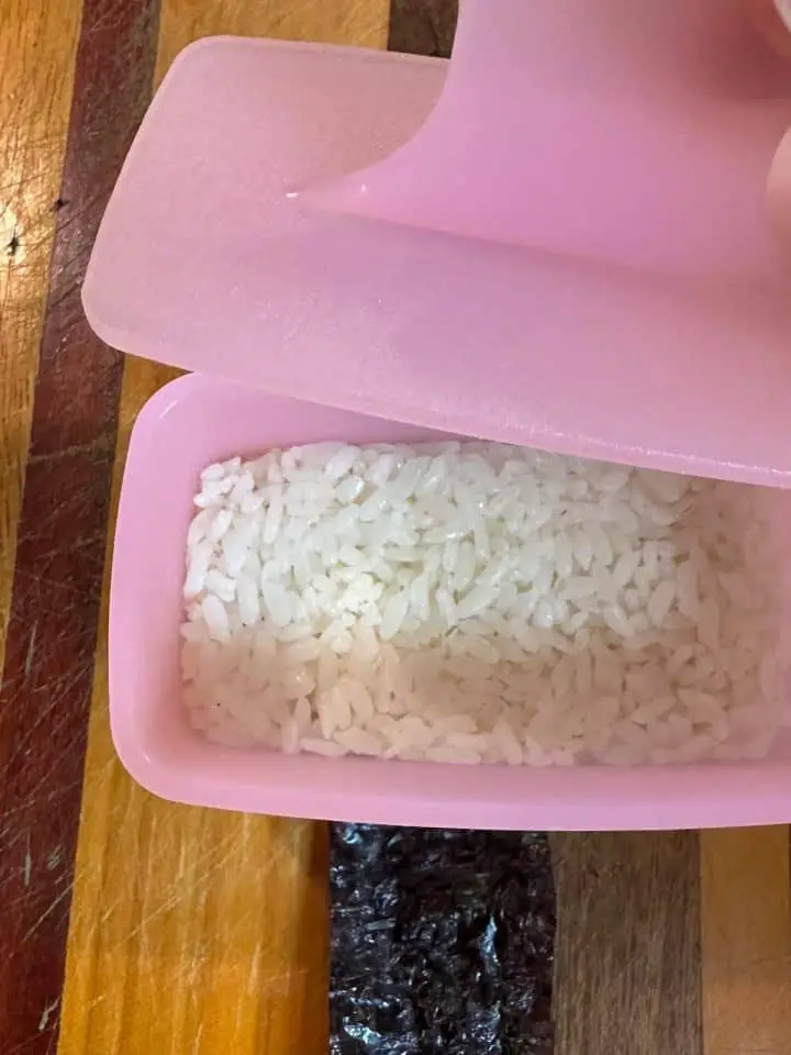 A strip of nori topped with a pink musubi maker. There is a bed of rice in the musubi maker and there is the presser part of the musubi maker poised over the bed of rice ready to press it down.