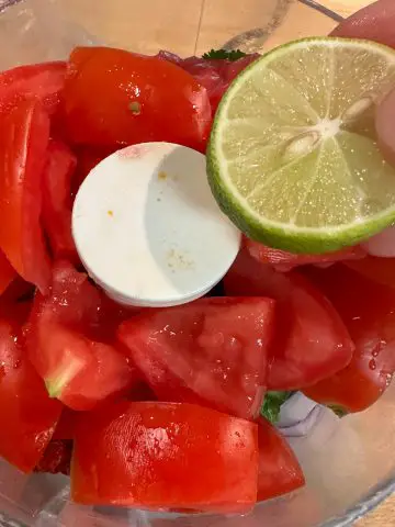 A food processor containing Ghost pepper salsa ingredients including chopped tomatoes. Someone is holding a slice of key lime over the food processor.