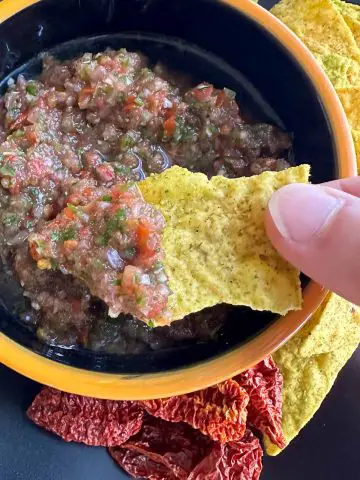 Ghost pepper salsa in a bowl surrounded by tortilla chips and dried Ghost chilies. There is someone holding a tortilla chip which had been dipped into the salsa.