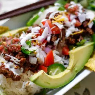 Japanese Taco rice which is rice topped with taco seasoned ground beef garnished with avocado, tortilla chips, diced tomato and onion, sliced jalapenos, shredded lettuce and cheese, sour cream and salsa in a white dish with chopsticks resting on the side of the dish.