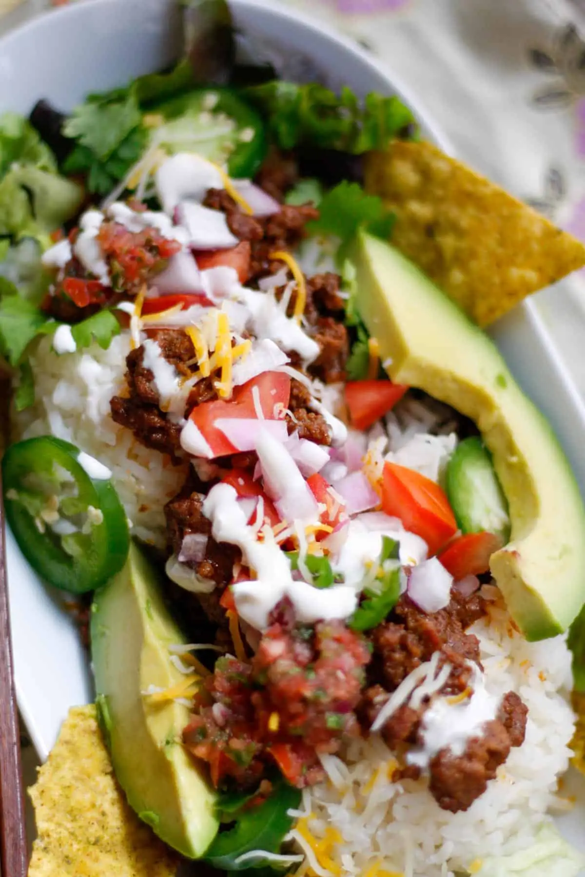 Japanese Taco rice which is rice topped with taco seasoned ground beef garnished with avocado, tortilla chips, diced tomato and onion, sliced jalapenos, shredded lettuce and cheese, sour cream and salsa in a white dish.