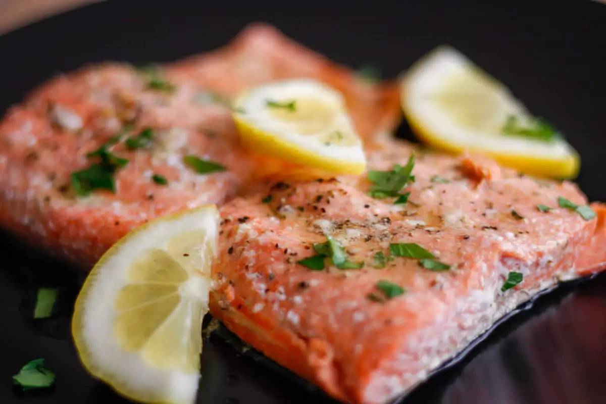 Cooked salmon fillets seasoned with salt and pepper, and garnished with lemon wedges and Italian parsley.