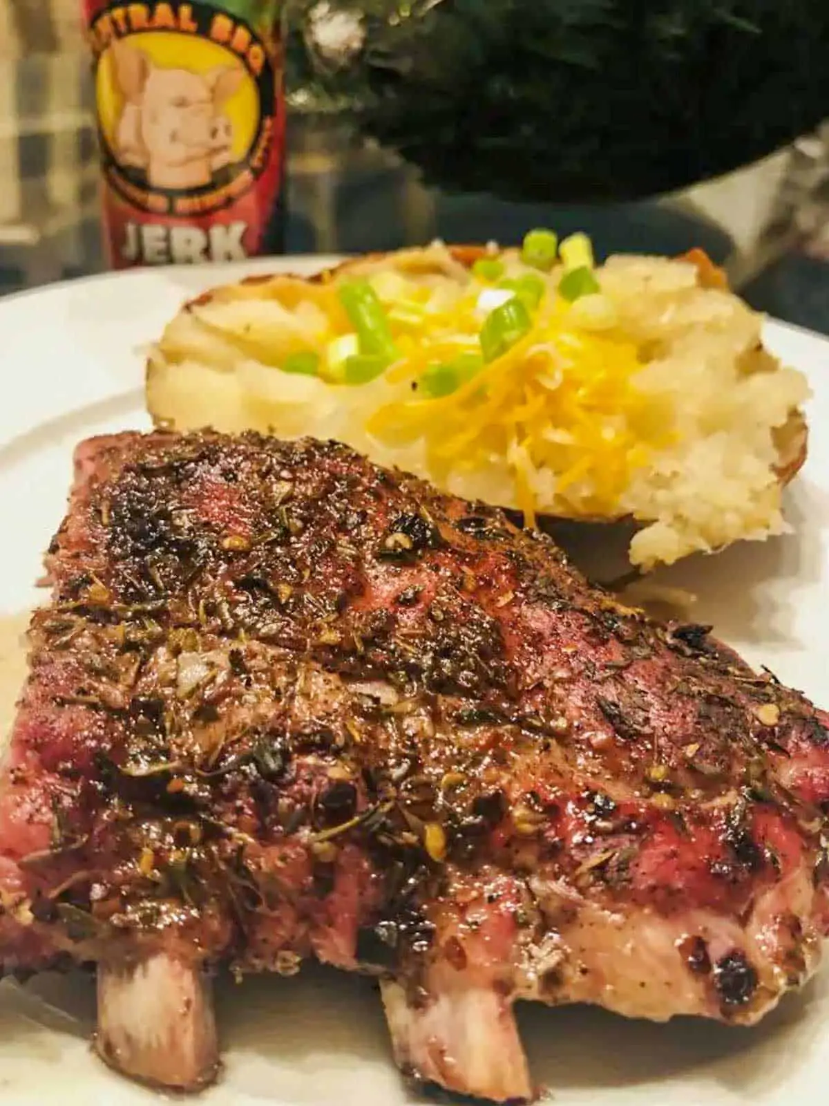 A rack of baby back pork ribs and a baked potato with cheese and green onions on a white plate. There is a bottle of Central BBQ Jerk Seasoning in the background.