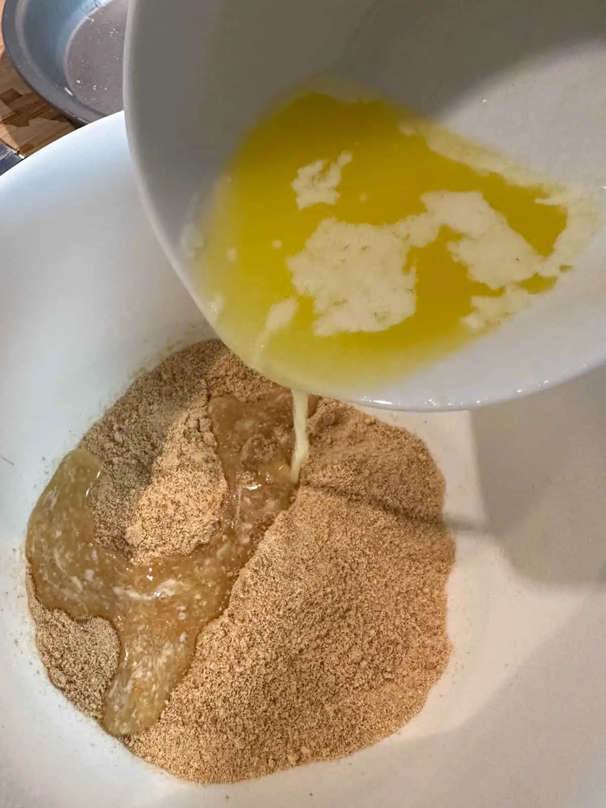 A mixing bowl containing crushed graham crackers. There is a white bowl containing melted butter that is being poured into the mixing bowl.