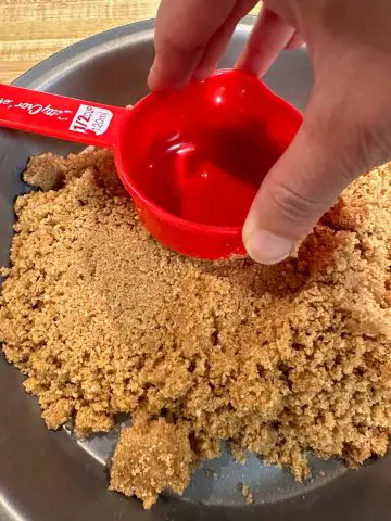 A pie pan with crushed graham crackers. There is a person holding a red measuring cup and pressing it down on the graham crackers.