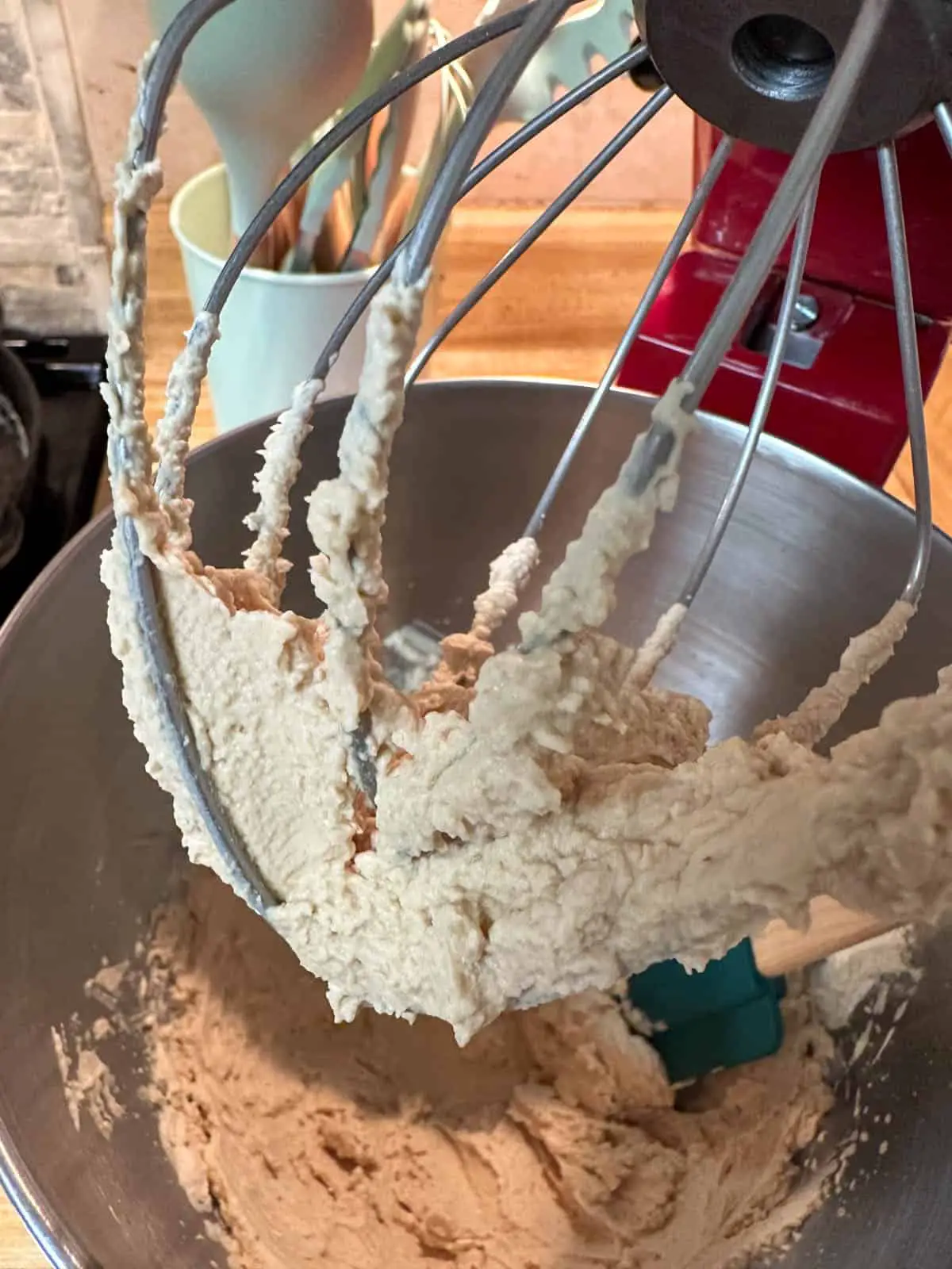 A KitchenAid stand mixer with whipped cream in the mixing bowl and on the whisk attachment. There is a set of kitchen utensils in the background.