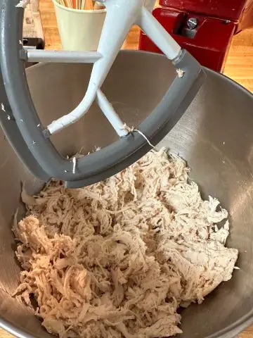 A stand mixer bowl with shredded chicken in it. The stand mixer has the flat beater attached to it.