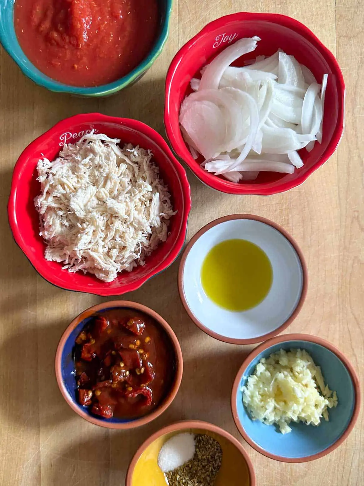 Bowls containing crushed tomatoes, sliced white onions, shredded chicken, chopped chipotle chilies in adobo sauce, olive oil, minced garlic, and seasonings.