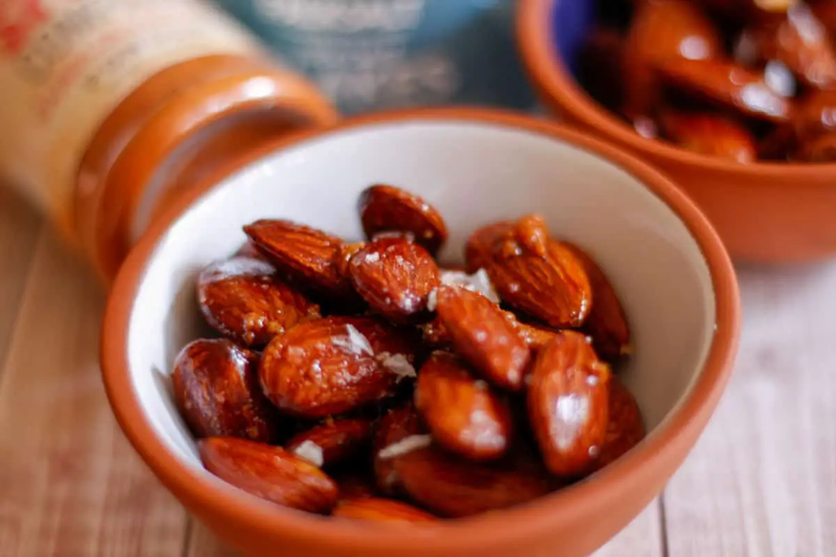 Small bowls filled with roasted almonds garnished with sea salt flakes, and a container of sea salt flakes and bottle of Carolina Reaper powder in the background.