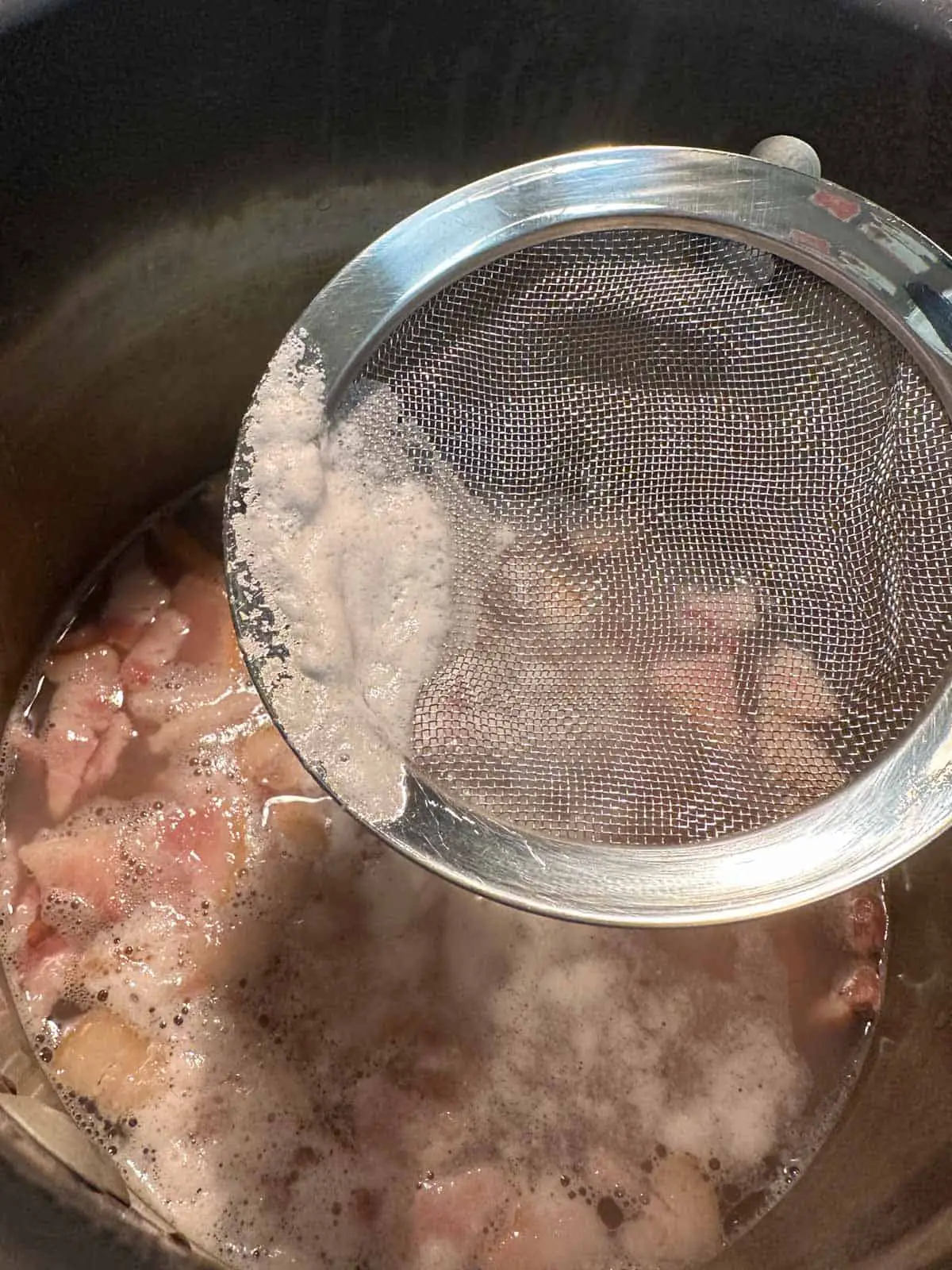 A large pot containing water, bacon, pinto beans, and scum. There is a strainer spoon over the pot containing some scum scooped up from the top of the broth.