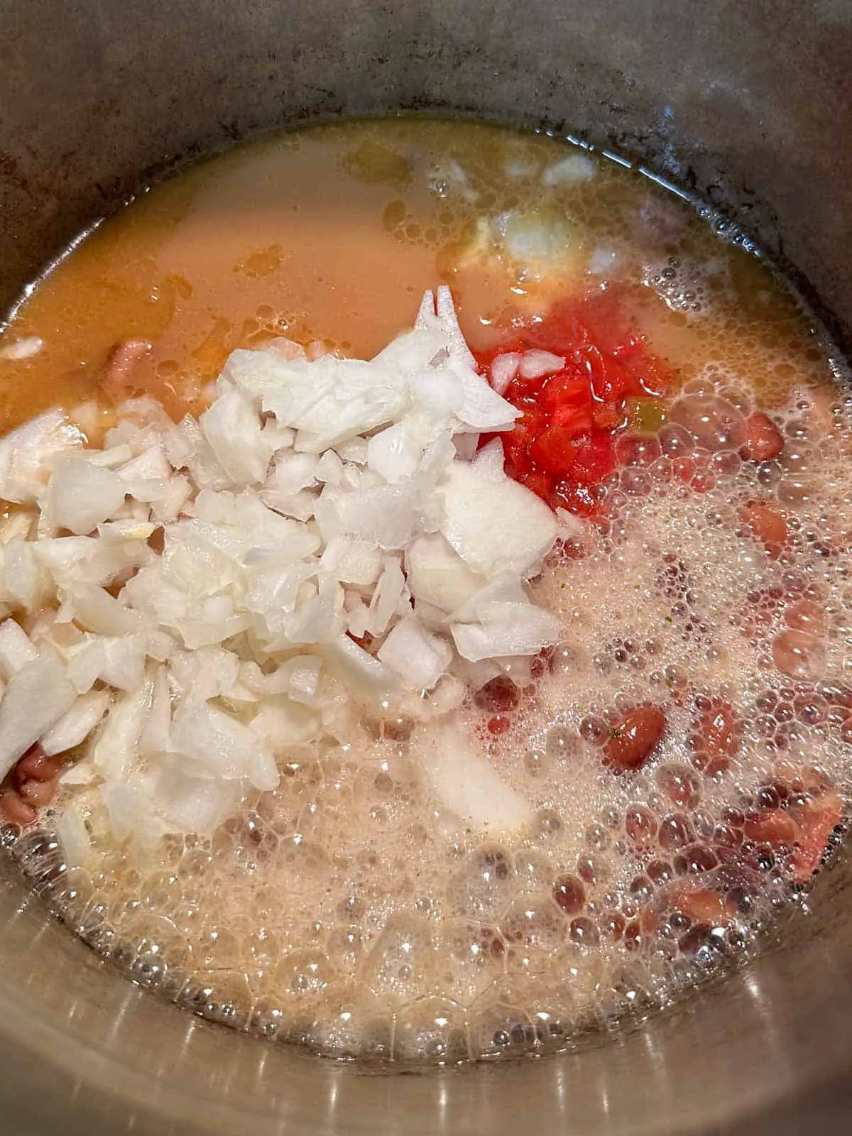 A large pot containing broth, beans, bacon, chopped onion, diced tomatoes, and beer.