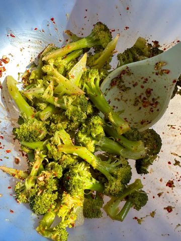 Broccoli florets dressed with a Korean style sauce which includes garlic and Korean red pepper flakes in a blue bowl. There is a blue spoon in the bowl.