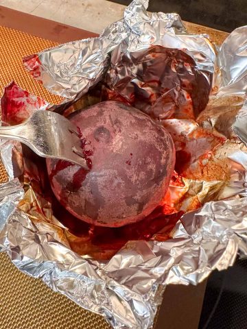 A beet pierced with a fork to show the beet is fork tender. The beet has released some of its juices and is in a piece of aluminum foil.