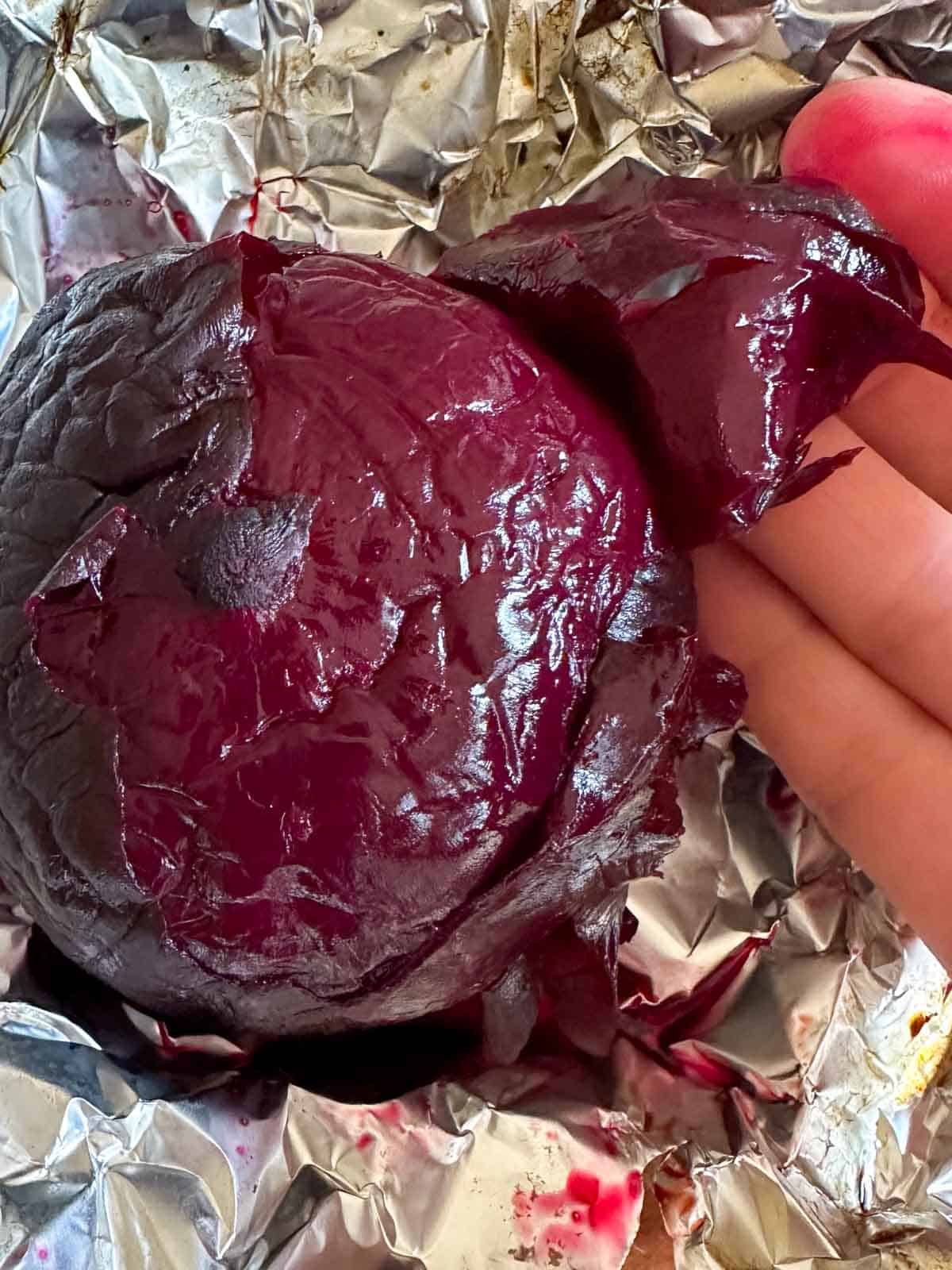 A whole beet that has been roasted in the oven on a sheet of aluminum foil, and a hand holding some of the skin of the beet which has been peeled off.