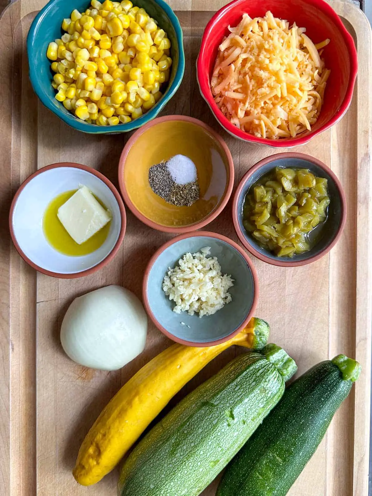 Bowls containing corn, shredded cheddar cheese, butter and olive oil, seasoning, green chilies and garlic. There are also half an onion and 3 different squashes pictured.