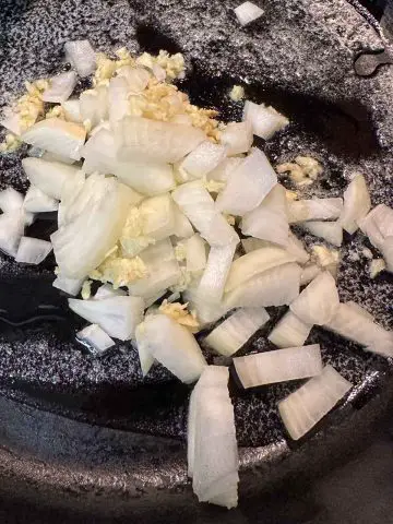 Chopped onion and minced garlic being cooked in butter and oil in a cast iron pan.