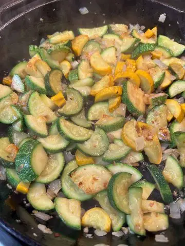 Sliced cooked squash and chopped onion being cooked in a cast iron pan.