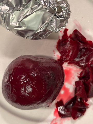 A foil wrapped beet and a peeled beet with the beet peels on a white plate.