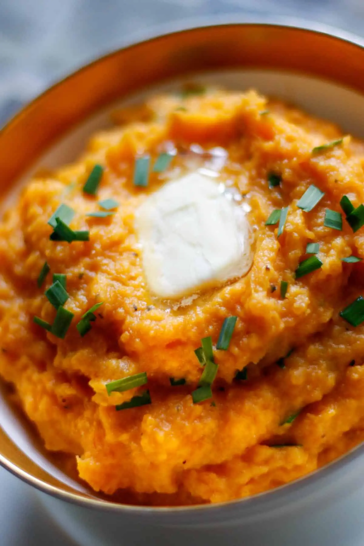 Sweet mashed potatoes in a gold rimmed bowl garnished with chopped chives with melting butter on the top.