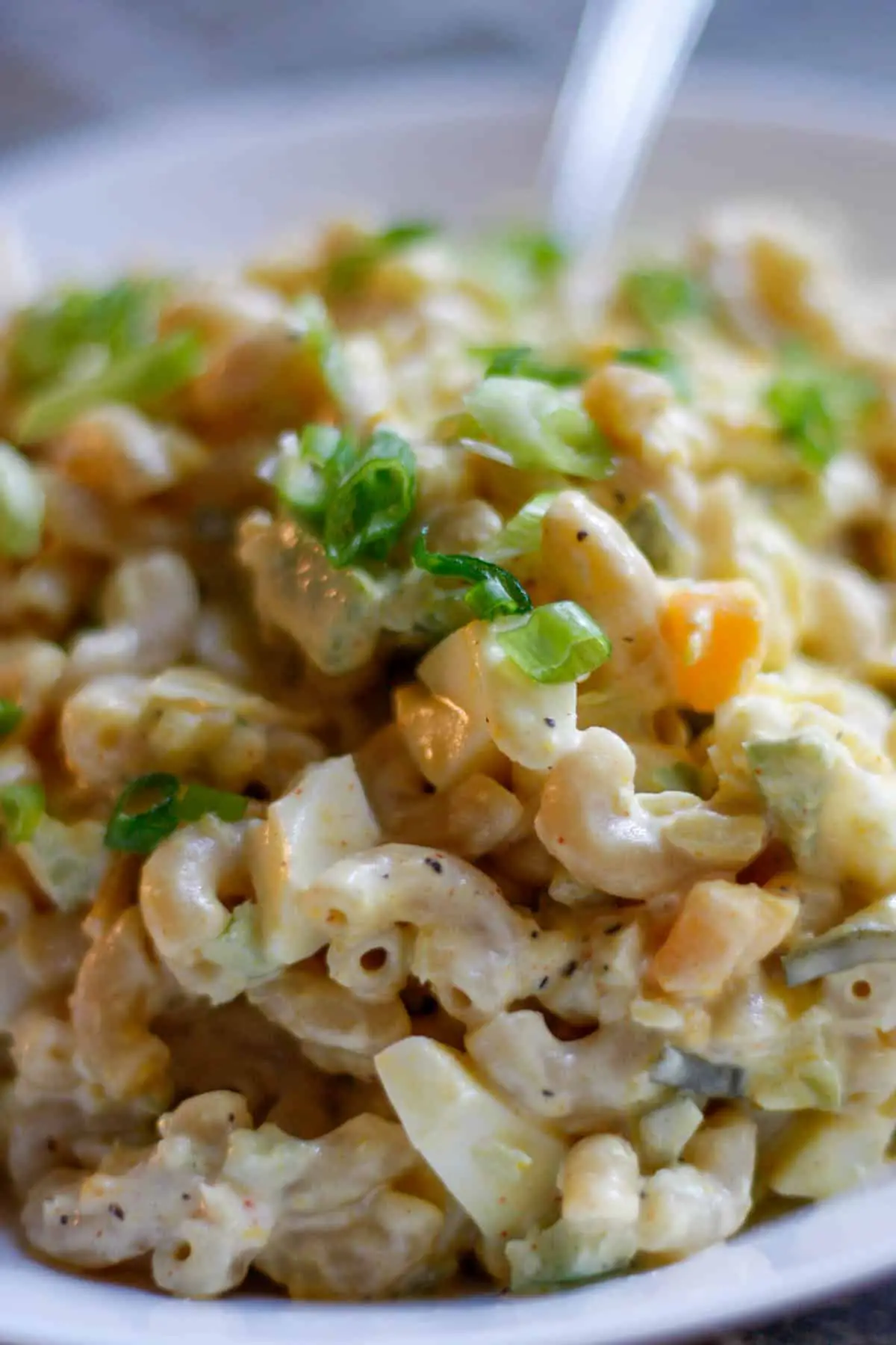 Southern macaroni salad with eggs and mayo garnished with green onions in a white dish, with the handle of a spoon visible in the background.