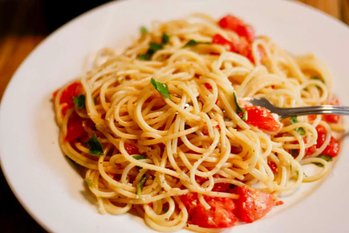 A white plate containing Pasta Alla Checca which is spaghetti with basil, tomatoes, and garlic. There is a fork inserted into the pasta.