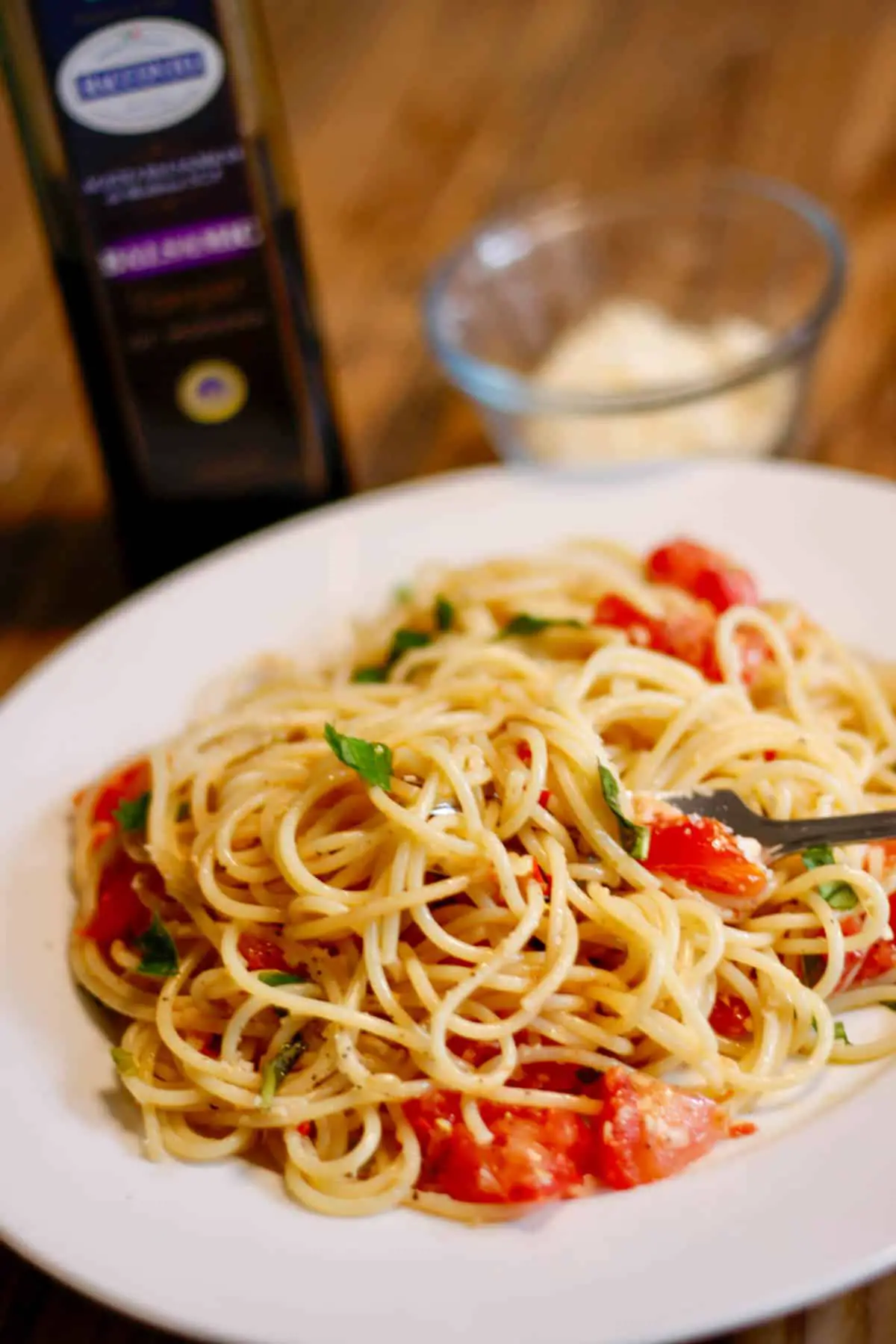 A white plate containing Pasta Alla Checca which is spaghetti with basil, tomatoes, and garlic. There is a fork inserted into the pasta and a bottle of Balsamic vinegar and bowl with Parmesan in the background.