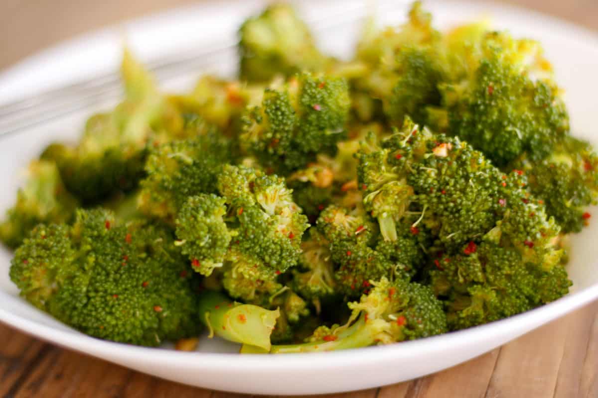 Broccoli florets seasoned in a Korean style sauce and flecked with red pepper flakes in a white bowl. There is a pair of silver chopsticks resting on the bowl in the background.