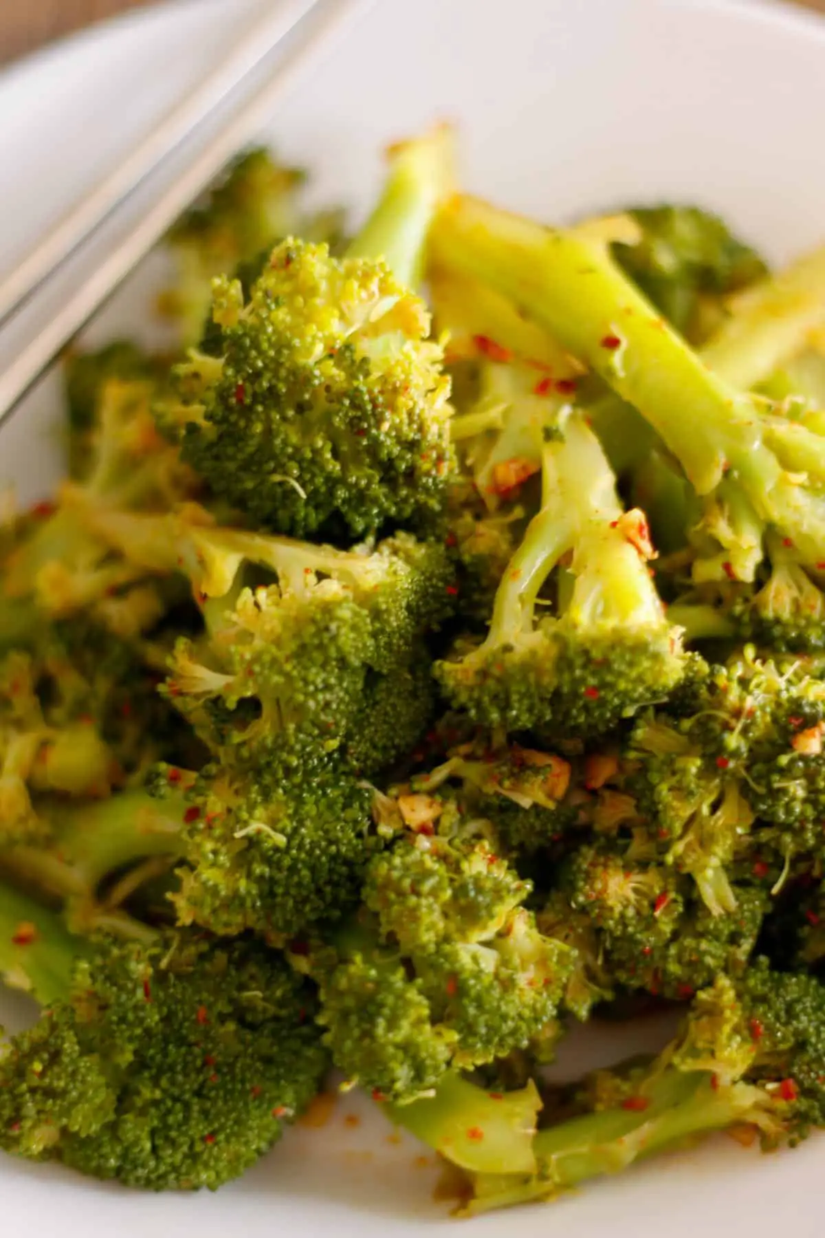 Broccoli florets seasoned in a Korean style sauce and flecked with red pepper flakes in a white bowl. There is a pair of silver chopsticks resting on the bowl in the background.