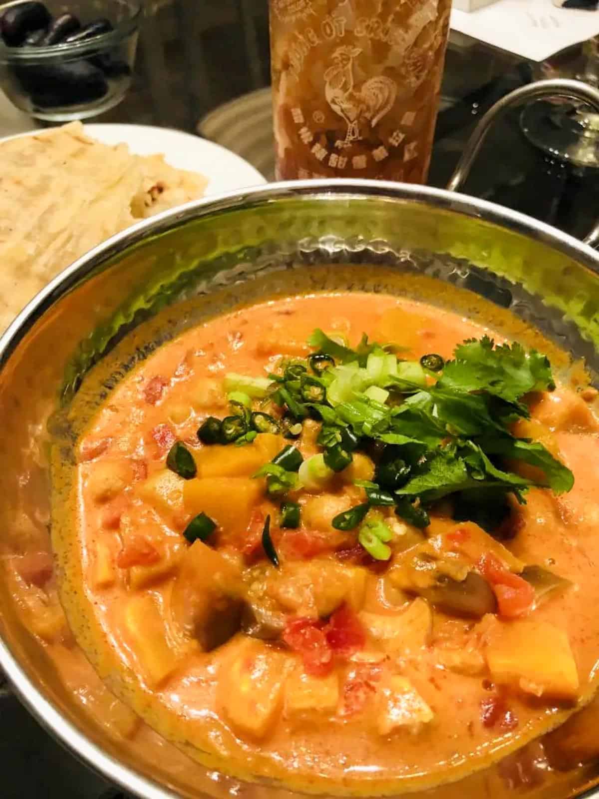 Thai Panang Curry with vegetables garnished with cilantro and green onions in a silver bowl. There is a plate with bread, bowl with olives, and bottle of hot sauce in the background.