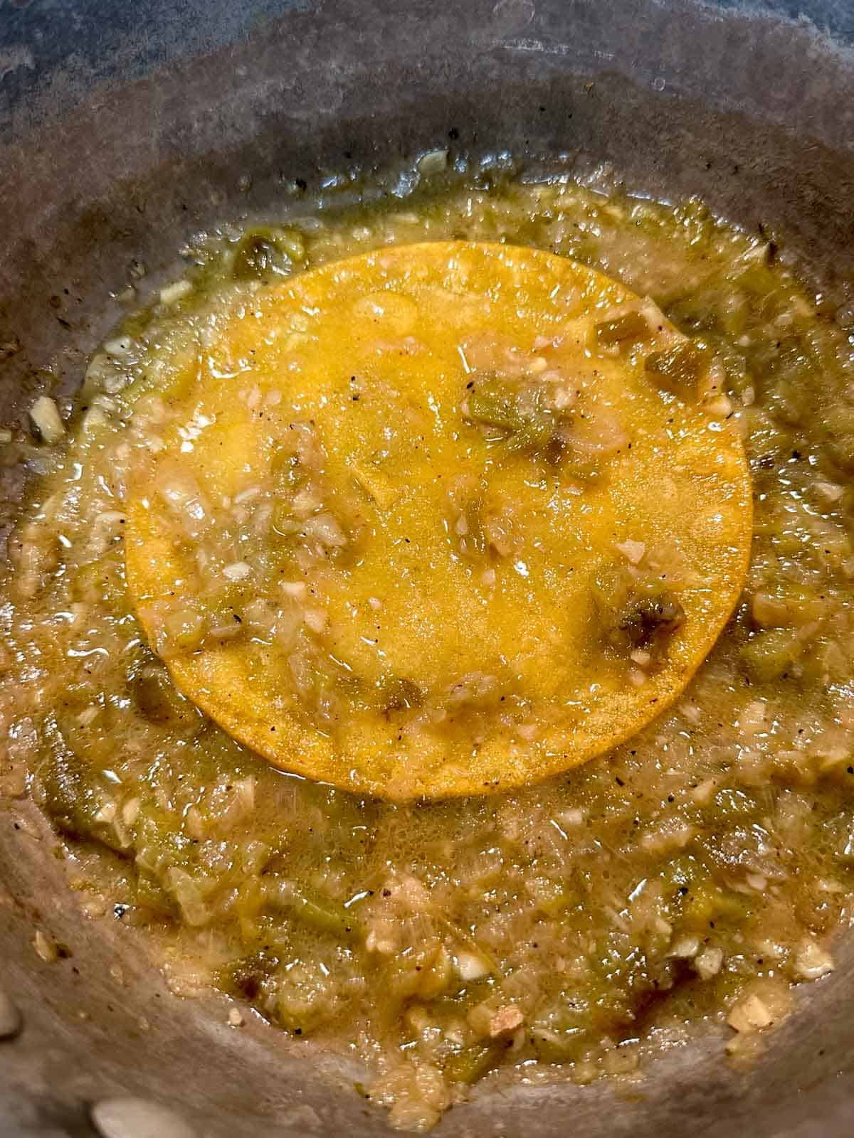 A corn tortilla sitting atop green chile sauce in a saucepan.