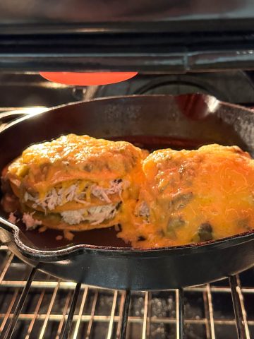 A cast iron skillet under a broiler containing stacked green chile chicken enchiladas smothered with melted cheese.