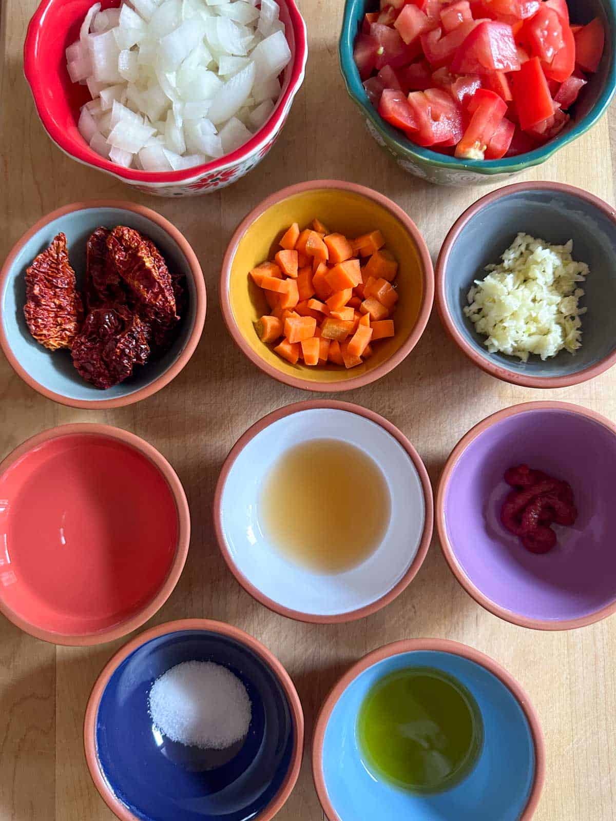 Bowls containing chopped onion, chopped tomatoes, dried ghost peppers, chopped carrot, minced onion, water, apple cider vinegar, tomato paste, salt, and olive oil.