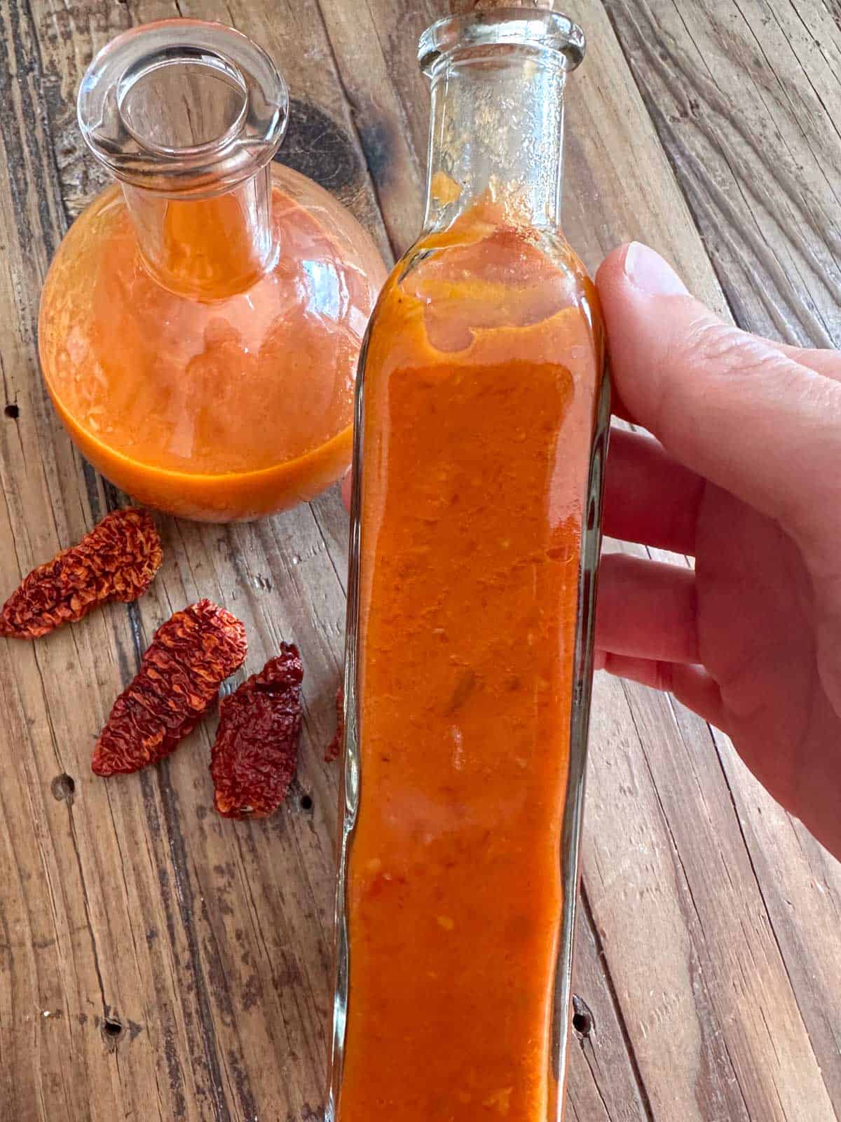 Glass bottles containing homemade ghost pepper hot sauce and some dried ghost peppers by the glass bottles and a person is holding one of the glass bottles.