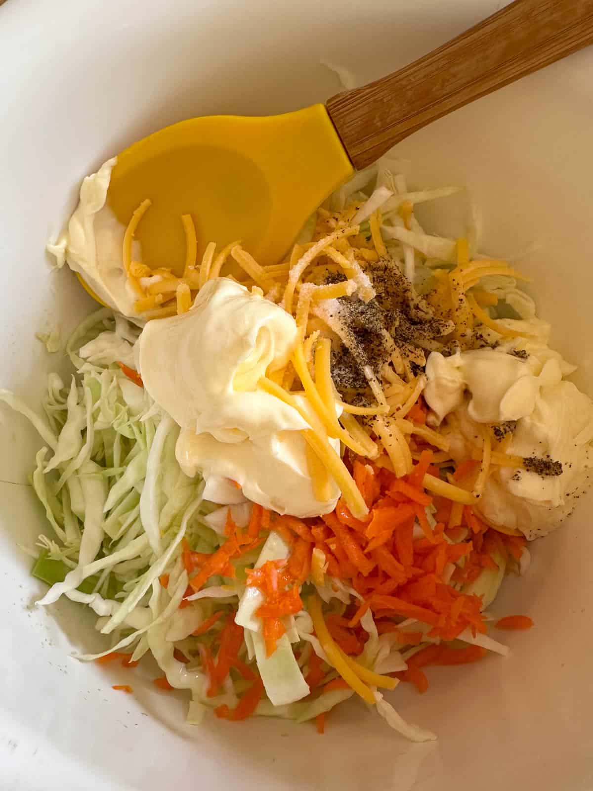 A mixing bowl containing the coleslaw ingredients including shredded cabbage and carrots, mayonnaise, and salt and pepper. There is a yellow spoon with a wooden handle in the bowl.
