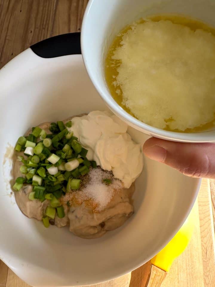 A white mixing bowl containing sour cream, cream of mushroom soup, green onions and seasonings. There is a white bowl with melted butter poised above the mixing bowl.