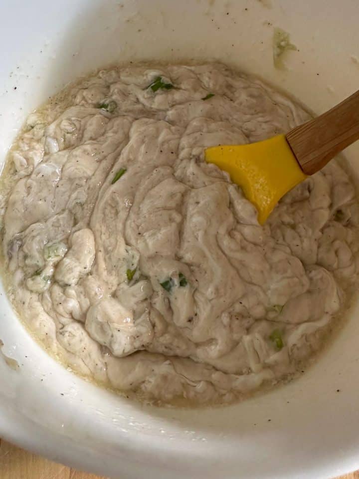 A mixing bowl containing a liquid mixture of cream of mushroom soup, green onions, and sour cream. There is a yellow spoon with a wooden handle resting in the bowl.
